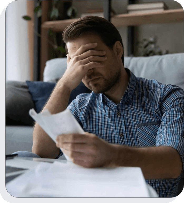 A man in a plaid shirt sits on a couch, covering his face with one hand while holding papers in the other, appearing stressed or frustrated. A table, laptop, and shelves are visible in the background.