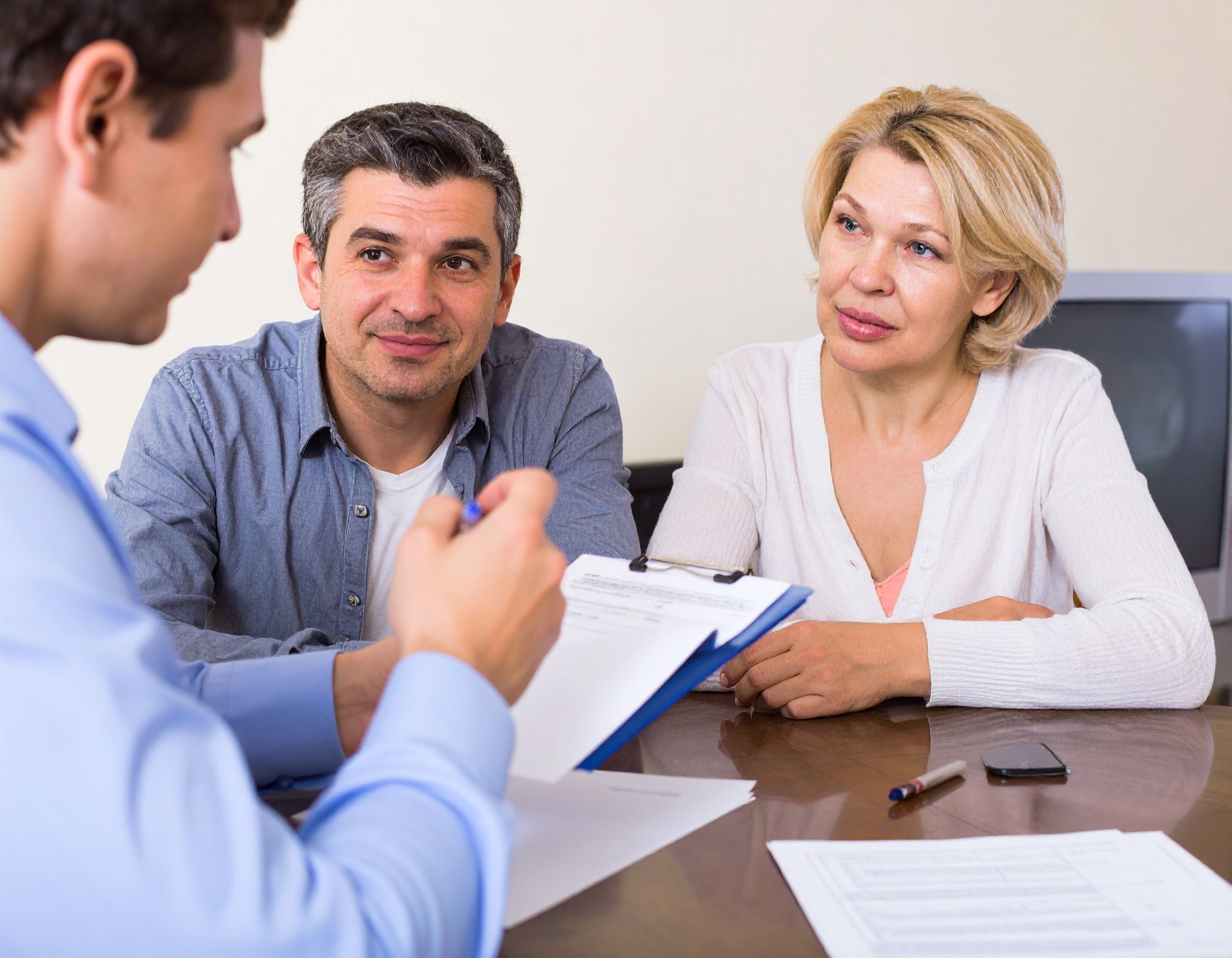 A man and woman sit at a table across from another man holding a clipboard. Papers, a pen, and a smartphone are on the table as the couple listens attentively to his advice about back taxes and possible tax resolution strategies.